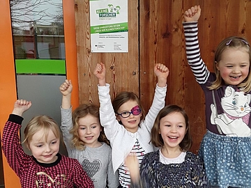 Jubelnde Kinder vor dem Kindergarten mit der Plakette „Haus der kleinen Forscher“ 